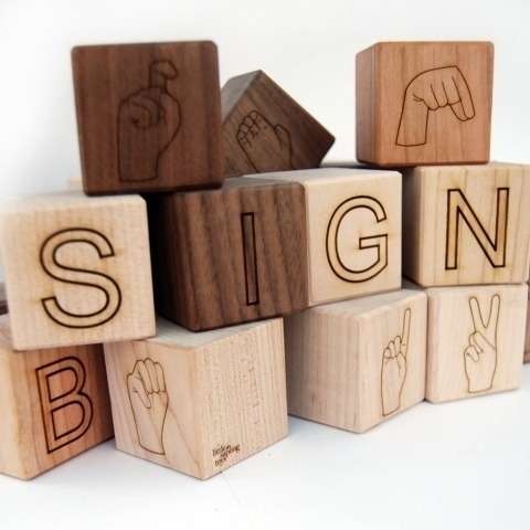 Wooden blocks in dark and light shades of wood spell out the word "SIGN" and depict several different letters in ASL.