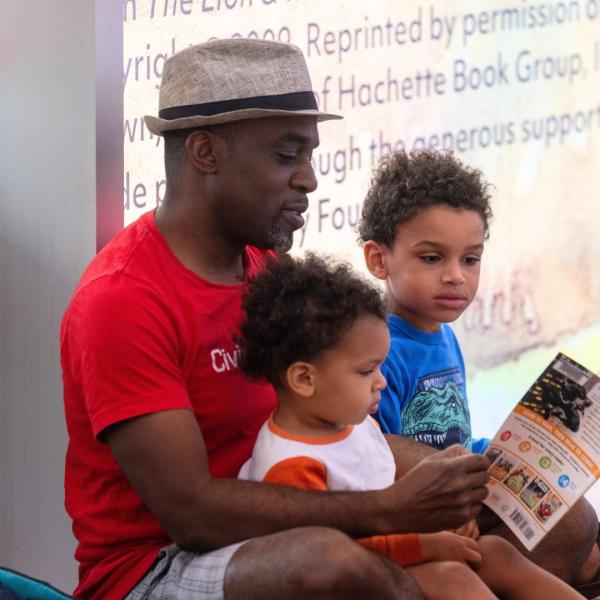 A man with two children in his lap, all three are looking at a book together.