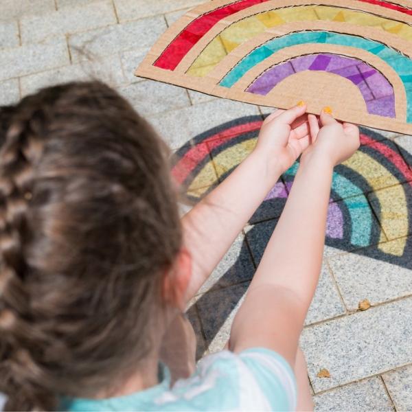 little girl holds a rainbow cardboard and plastic craft over the sidewalk looking at its reflection