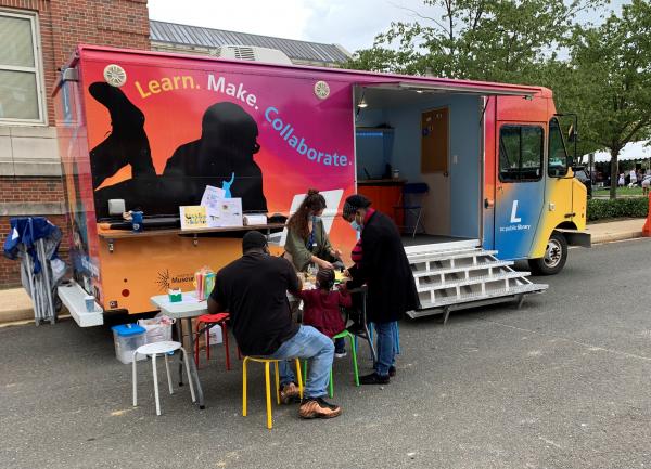 Photo of the brightly-colored Library on the Go-Go truck with a library staff person and three customers interacting outside.