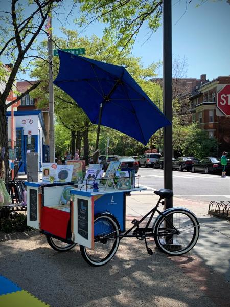 Open cargo bike with a blue umbrella displaying books on a shady street corner.