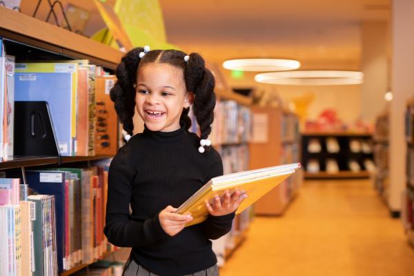 little girl stand among book shelves holding a book 
