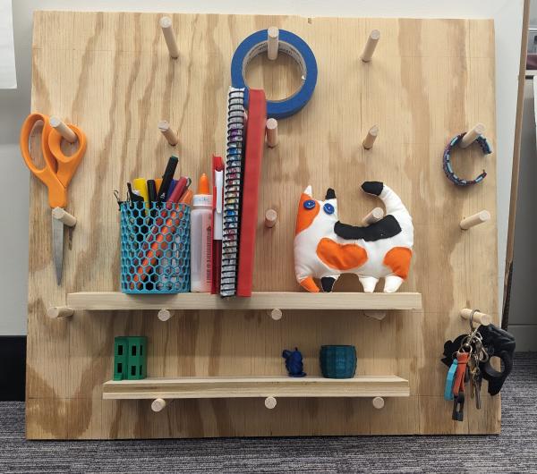 Peg board made out of scrap wood holding various object like: scissors, tin of pencils, notebooks, and a stuffed animal cat