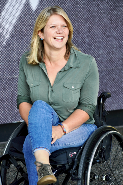 Photograph of Opie and the Open Tent creator, Regan Linton, a woman with straight blond hair, a button up green shirt, blue jeans, and tennis shoes, sitting in a wheelchair.