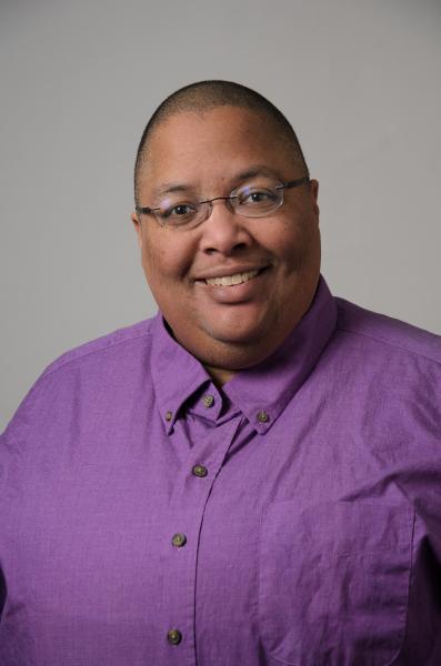 A picture of Tolonda Henderson, a smiling African American nonbinary person wearing a purple shirt and glasses