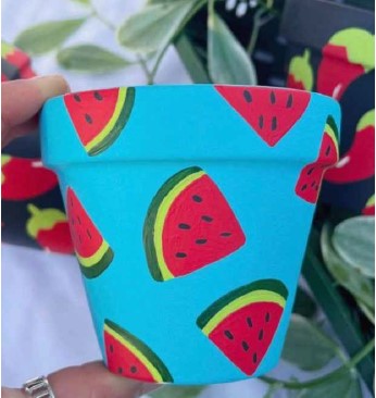 Photo of a small planter being held by index finger and thumb. The planter is painter with a turquoise blue background and slices of watermelons--with rinds of yellow and green, and red melon with black seeds-- around the planter.