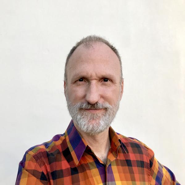 Photo of poet Raymond Luczak, a white man with a gray beard wearing an orange, purple, and yellow plaid shirt.