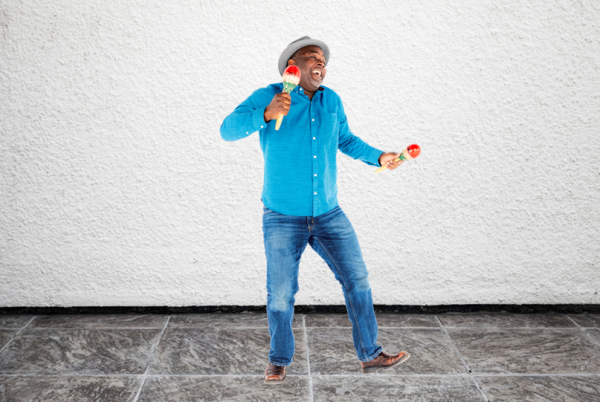 A smiling man in blue jeans, a blue button-up shirt, and a brimmed hat plays the the maracas and dances.